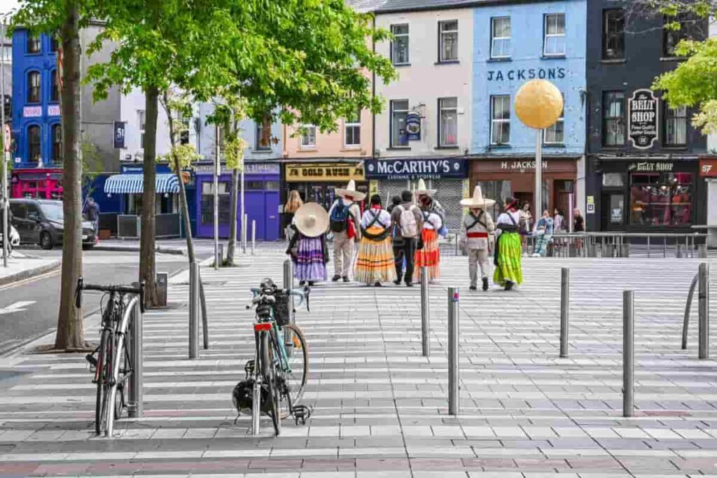 Group of people in colorful traditional costumes walking in an Irish town square with bicycles parked in the foreground.
