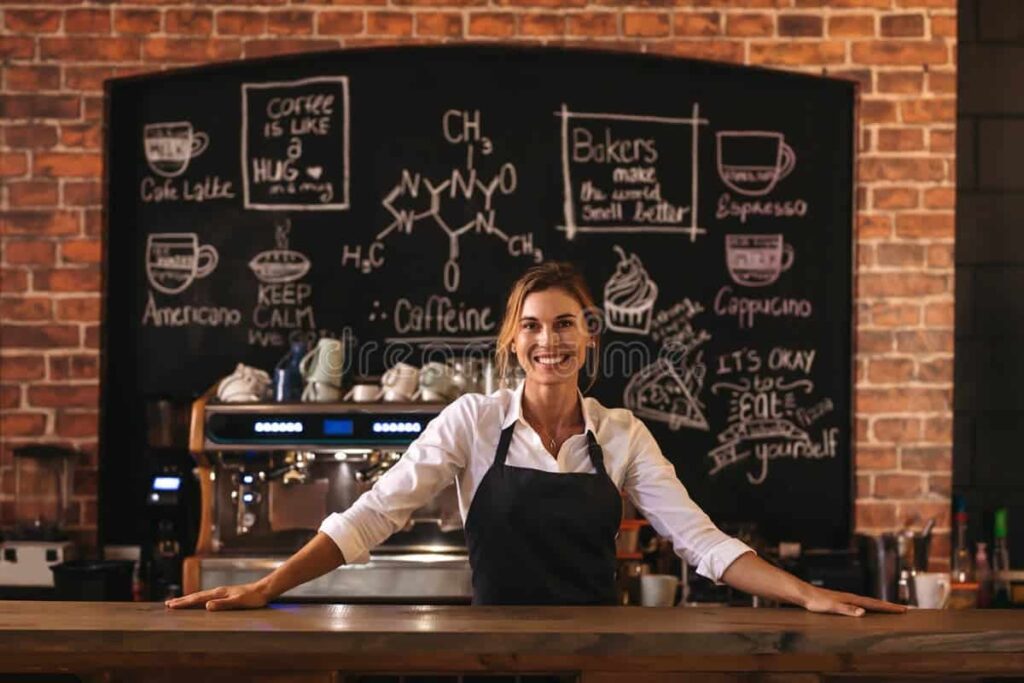 Smiling female barista standing behind the counter in a cozy coffee shop with chalkboard art.