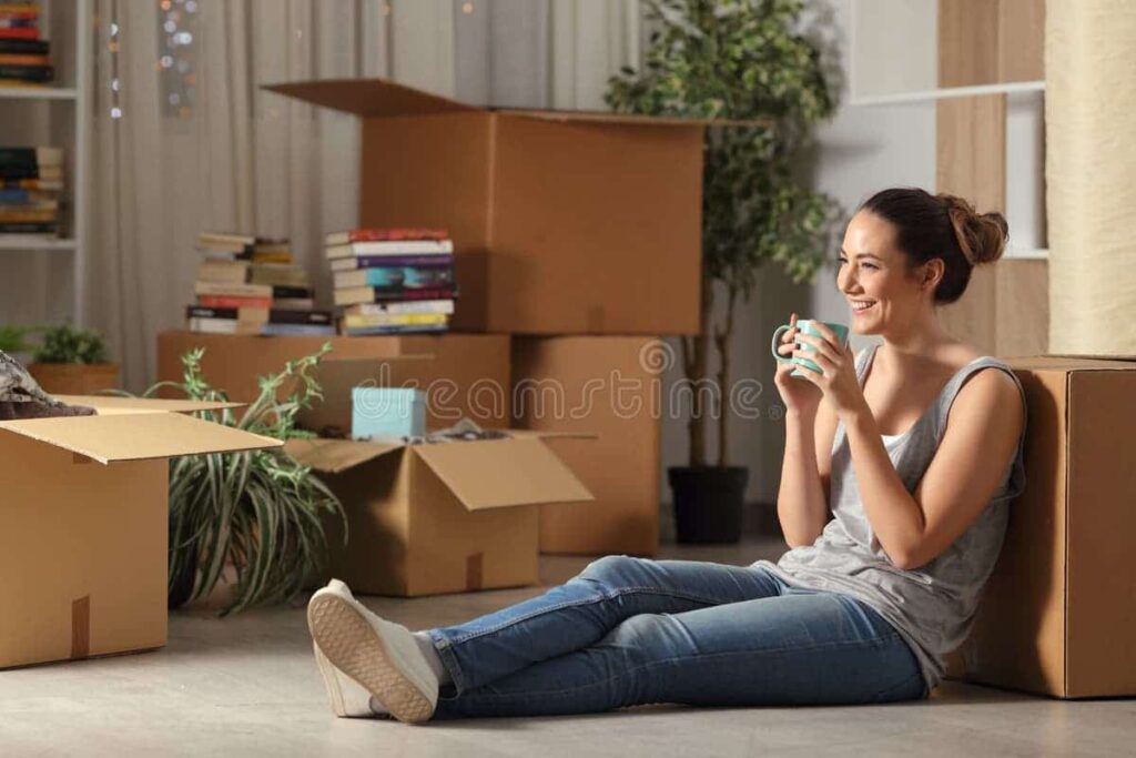 Young woman sitting on floor with coffee mug and moving boxes, symbolizing relocation or student accommodation setup.