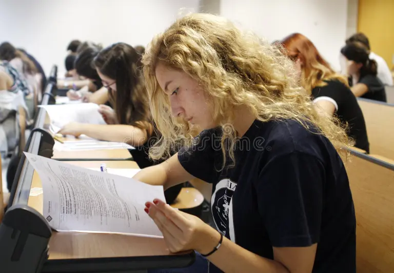 University student reading a handout in a lecture hall while classmates work on papers—exam or orientation setting.