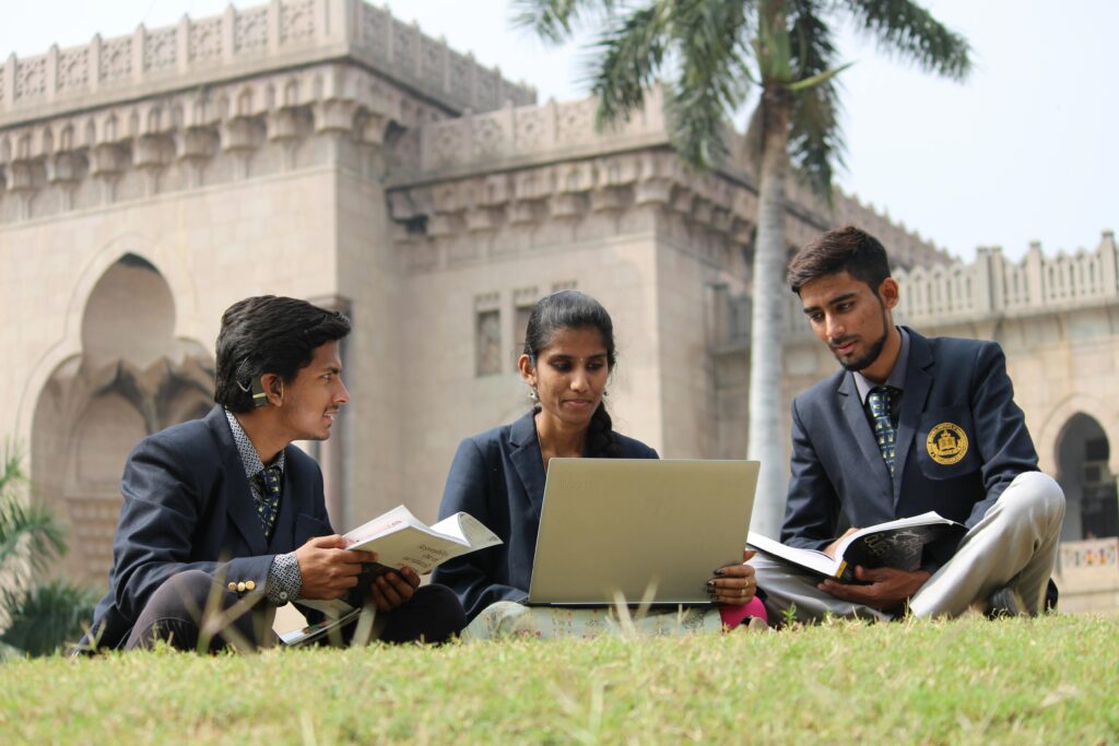 Three students studying with a laptop and books on a campus lawn in front of a heritage-style Indian university building.