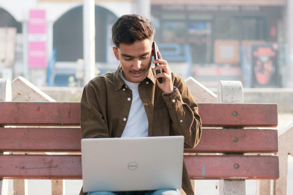 Student sitting on a bench using a laptop and talking on the phone, representing preparation for studying in India.