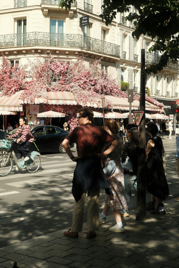 Paris street with people walking, a cyclist, and a beautifully decorated café with pink flowers, representing the cultural experience and lively lifestyle in France.