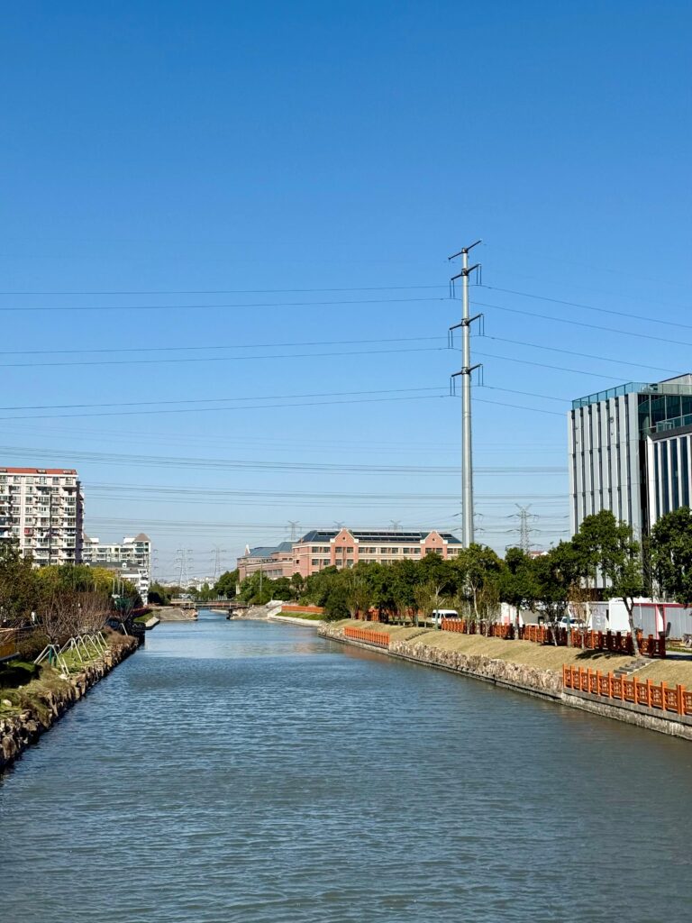 A calm river flowing between landscaped banks with trees and orange railings, surrounded by modern buildings and power lines under a clear blue sky.