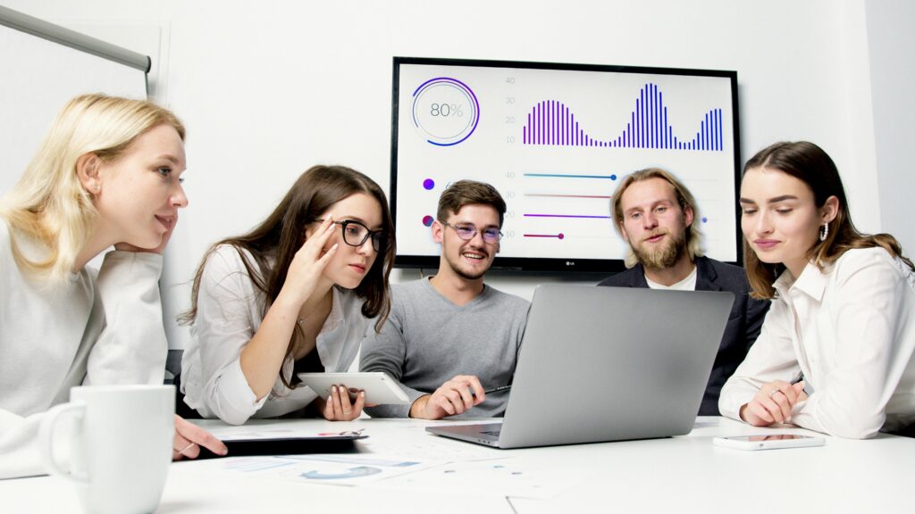 A group of young professionals sitting around a table, reviewing data on a laptop with analytics charts displayed on a screen behind them.