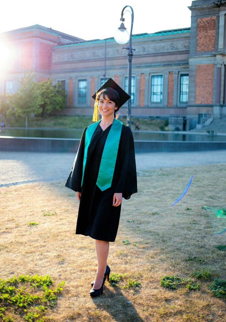 A smiling graduate wearing a cap and gown with a green stole, standing outdoors near a historic university building in Denmark.