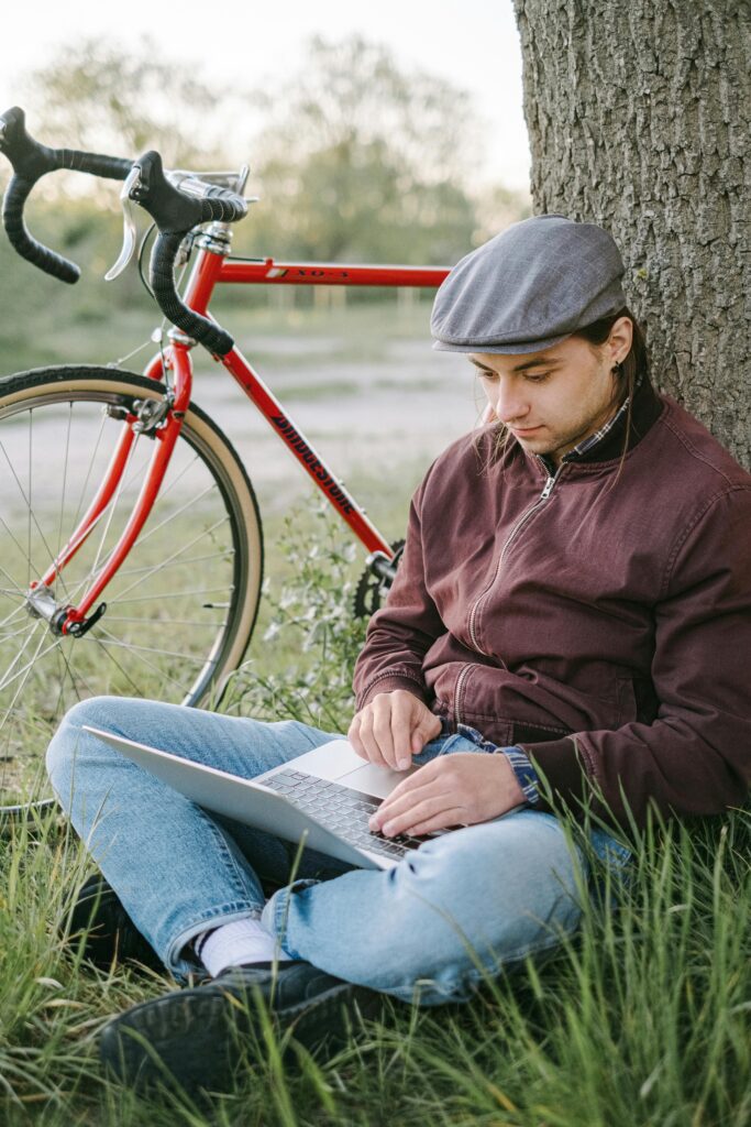 A student sitting under a tree with a bicycle nearby, working on a laptop outdoors — representing communication, learning, and connection for the Study Silently “Contact Us” section.