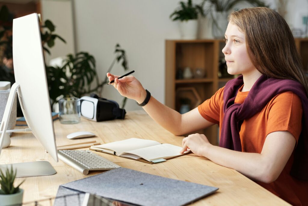 A focused student sitting at a desk, writing notes while looking at a computer screen in a bright study environment.