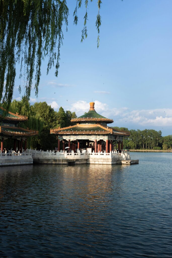 A traditional Chinese lakeside pavilion with green-tiled roofs, surrounded by calm water and hanging willow branches under a clear blue sky.