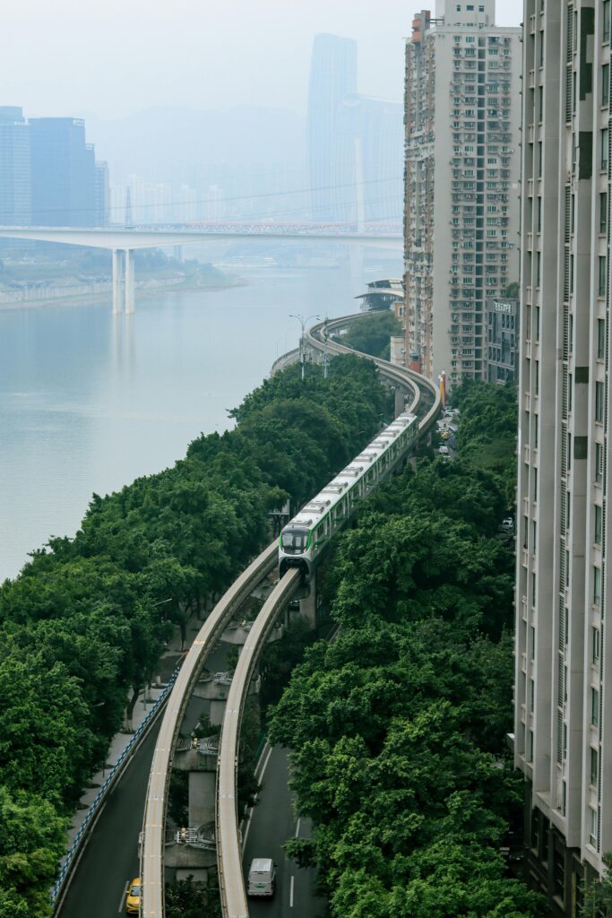 A modern monorail train traveling between high-rise buildings and dense greenery along a riverside in a Chinese city, symbolizing technology, money flow, and strong connectivity in China.