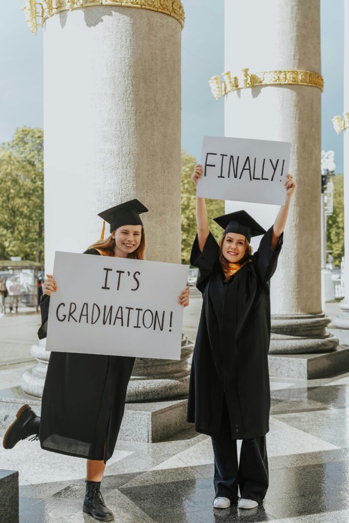 Two smiling graduates in black caps and gowns holding “It’s Graduation!” and “Finally!” signs while posing in front of tall columns.