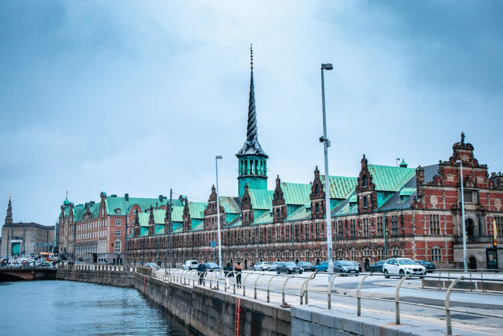 A scenic view of historic Danish buildings with green roofs beside a waterfront in a European city, under cloudy skies.