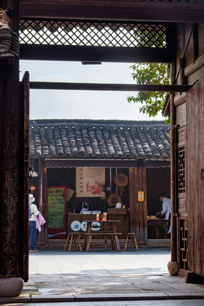 A view through a wooden gate into a traditional village shop with tiled roofs, wooden furniture, and people working inside.