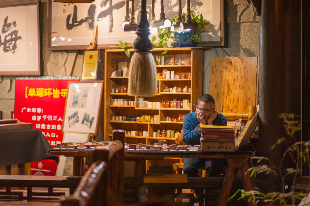 A Chinese man studying in a traditional calligraphy and seal-carving shop in China, surrounded by wooden shelves, brushes, books, and cultural art pieces. Warm lighting enhances the atmosphere of Chinese language and cultural learning.