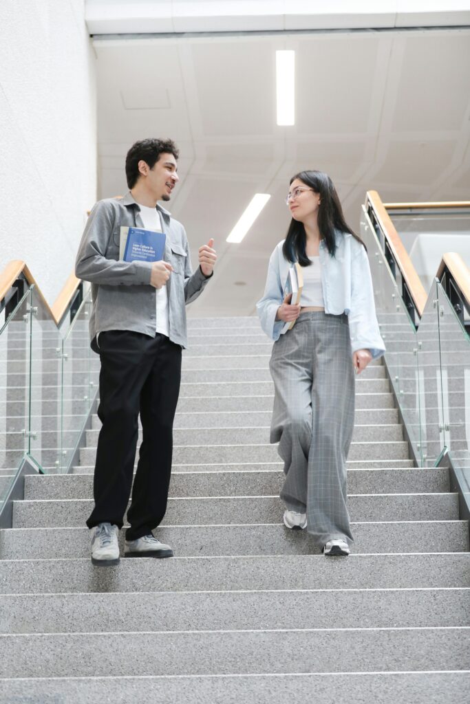 Two international university students walking down the stairs with books, discussing academics inside a modern educational campus in Singapore.
