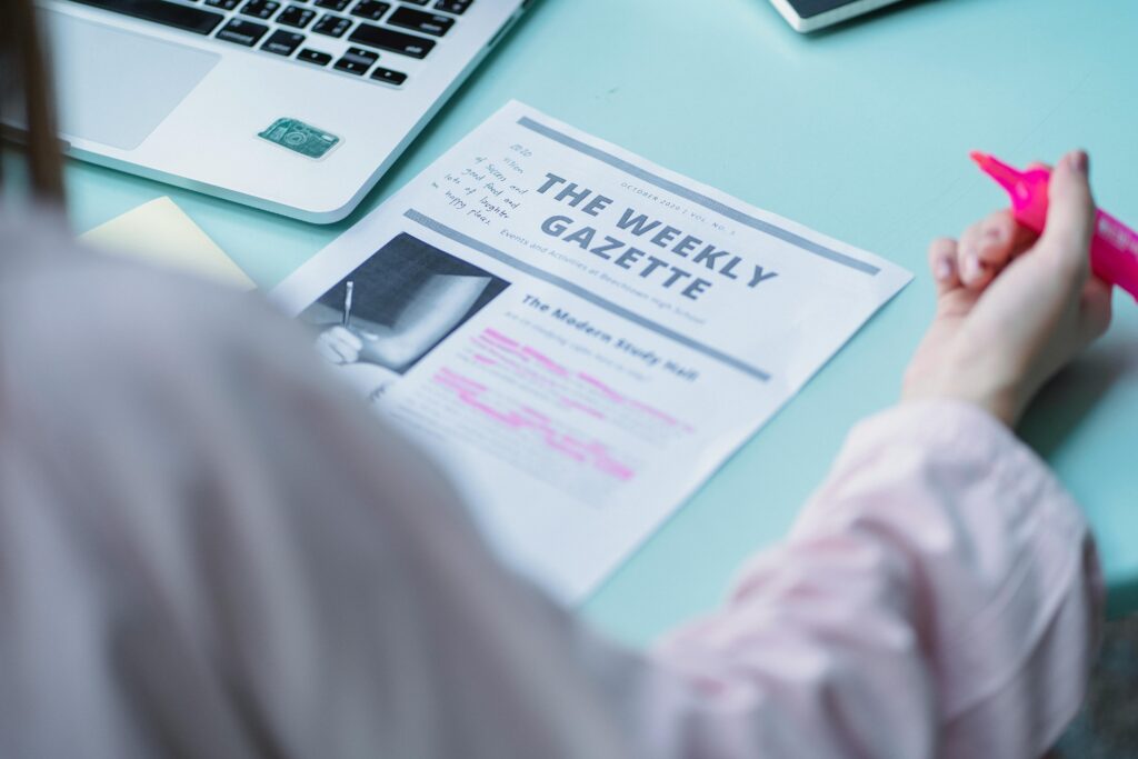 A person reading “The Weekly Gazette” newspaper beside a laptop, highlighting sections with a pink marker — representing browsing and engaging with the latest news and articles by category.